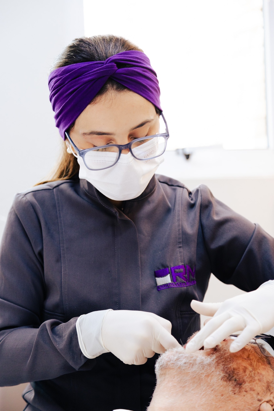 Dentista de máscara e luvas realizando procedimento cirúrgico em paciente. Profissional veste uniforme cinza com logo da RM Odontologia Integrativa na Vila Clementino.