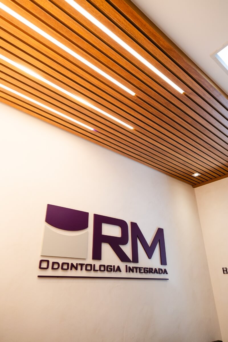 Logotipo da RM Odontologia Integrada fixado em parede branca sob teto de madeira ripada iluminado. Dentista na Vila Clementino.
