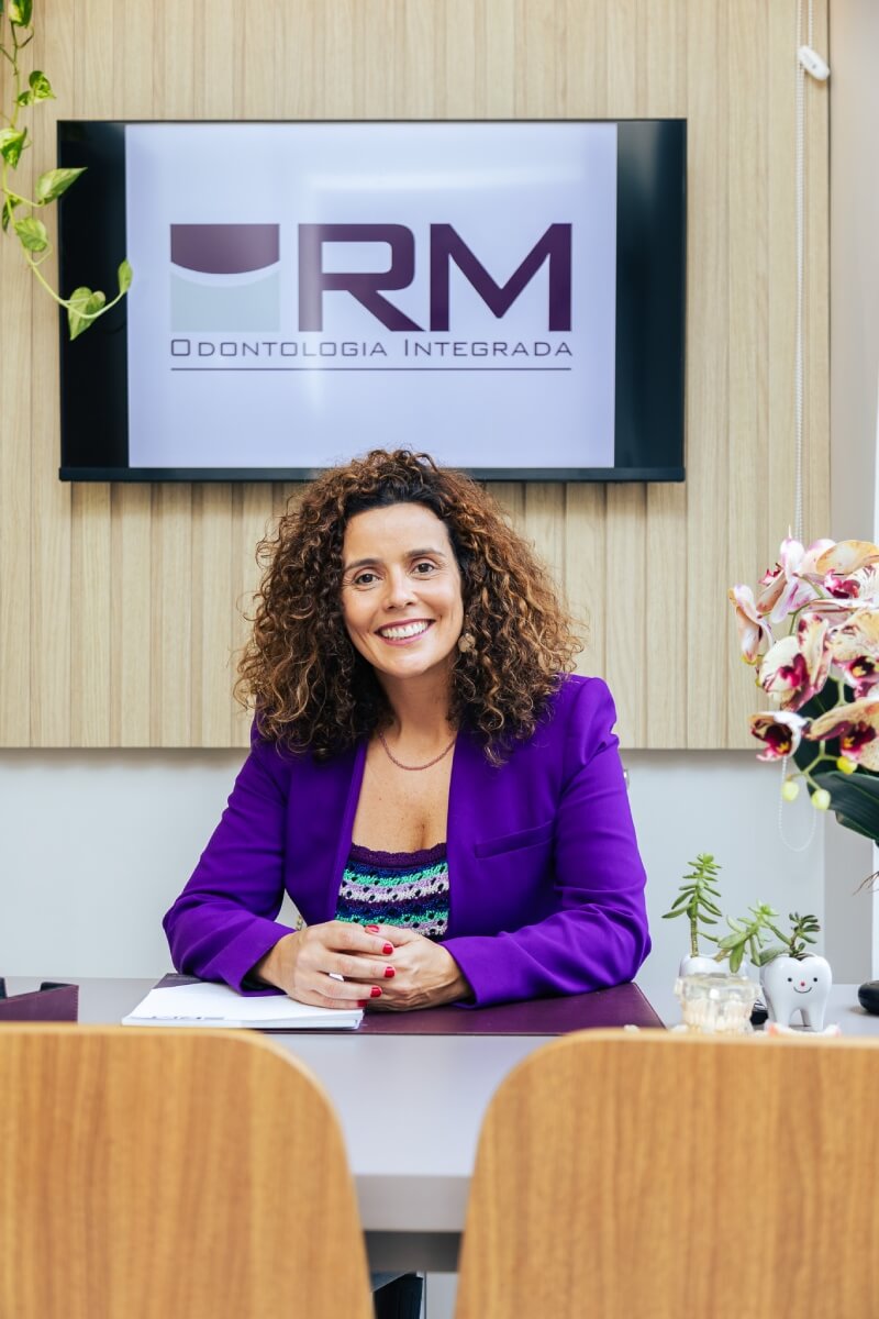 Dentista sorridente de blazer roxo sentada em seu consultório na Vila Clementino. Ao fundo, tela com logo da RM Odontologia Integrada e orquídeas. Se procura por