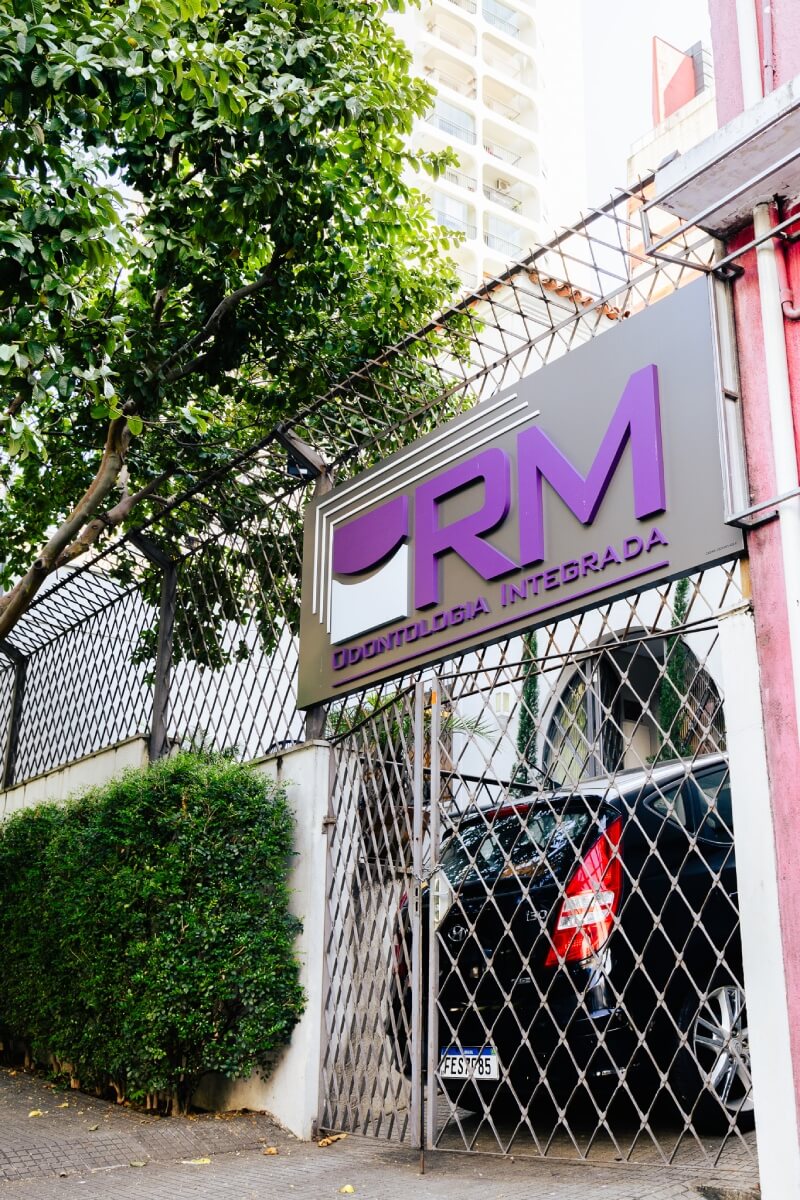 Fachada da clínica RM Odontologia Integrada com placa roxa em destaque, entrada com portão de ferro e jardim lateral. Dentista na Vila Clementino SP.